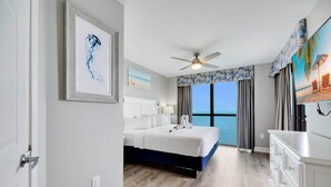 A modern hotel room with a large bed, a ceiling fan, a view of the ocean, and a white dresser.