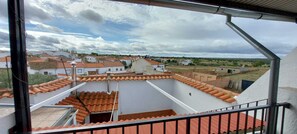 A view from a balcony with terracotta-tiled roofs, a white building, and a rural landscape in the background.