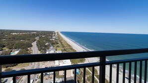 A coastal view from a balcony overlooking a beach and a road with houses and greenery.