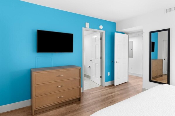 A bedroom with a bed, a dresser, a TV mounted on the wall, and a door leading to another room.