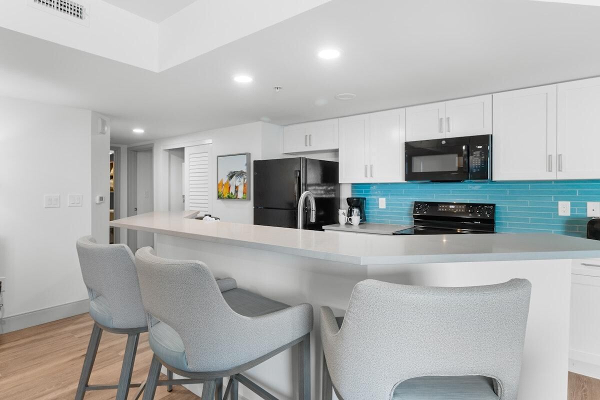 A modern kitchen with white cabinets, a black refrigerator, a microwave, and a blue tiled backsplash. There are three grey bar stools around the island.