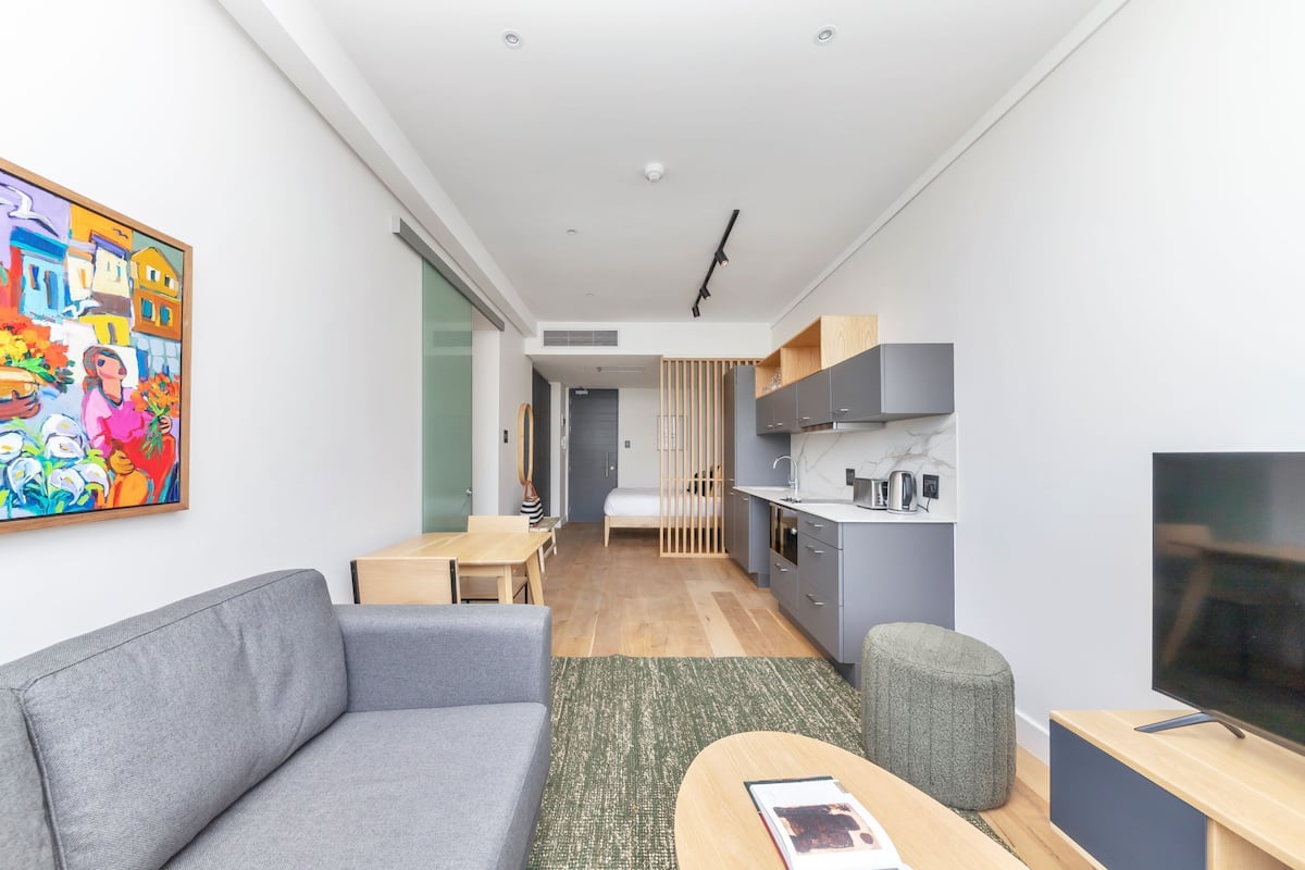 A modern apartment with a grey sofa, a small dining table, a kitchen area with grey cabinets, and a TV on a wooden stand.