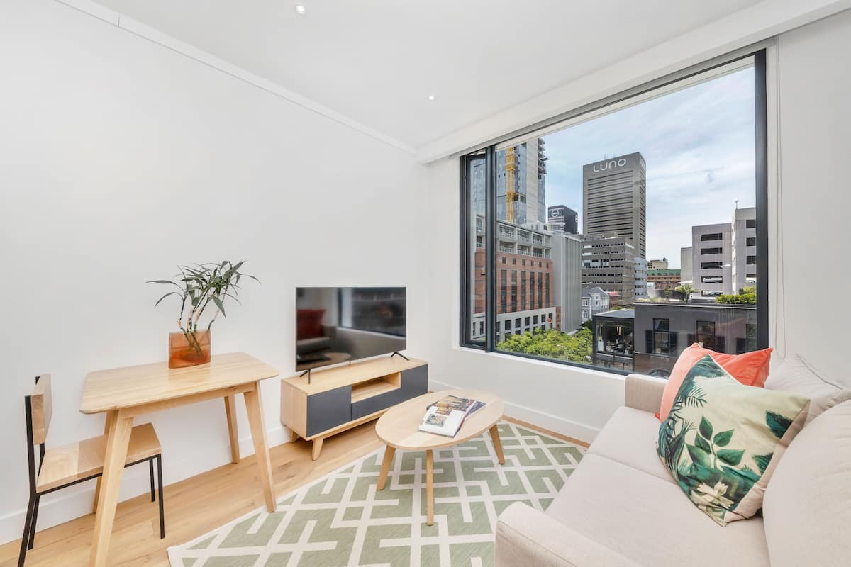A modern living room with a large window, a flat-screen TV, a wooden table, a sofa, and a patterned rug.