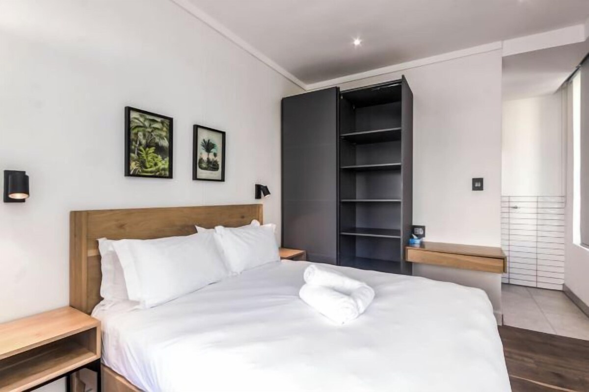 Ett modernt hotellrum med en sänggavel i trä, vita sängkläder, en prydligt bäddad säng med en handduk, ett litet träbord vid sängen, en hylla på väggen och två inramade tavlor.