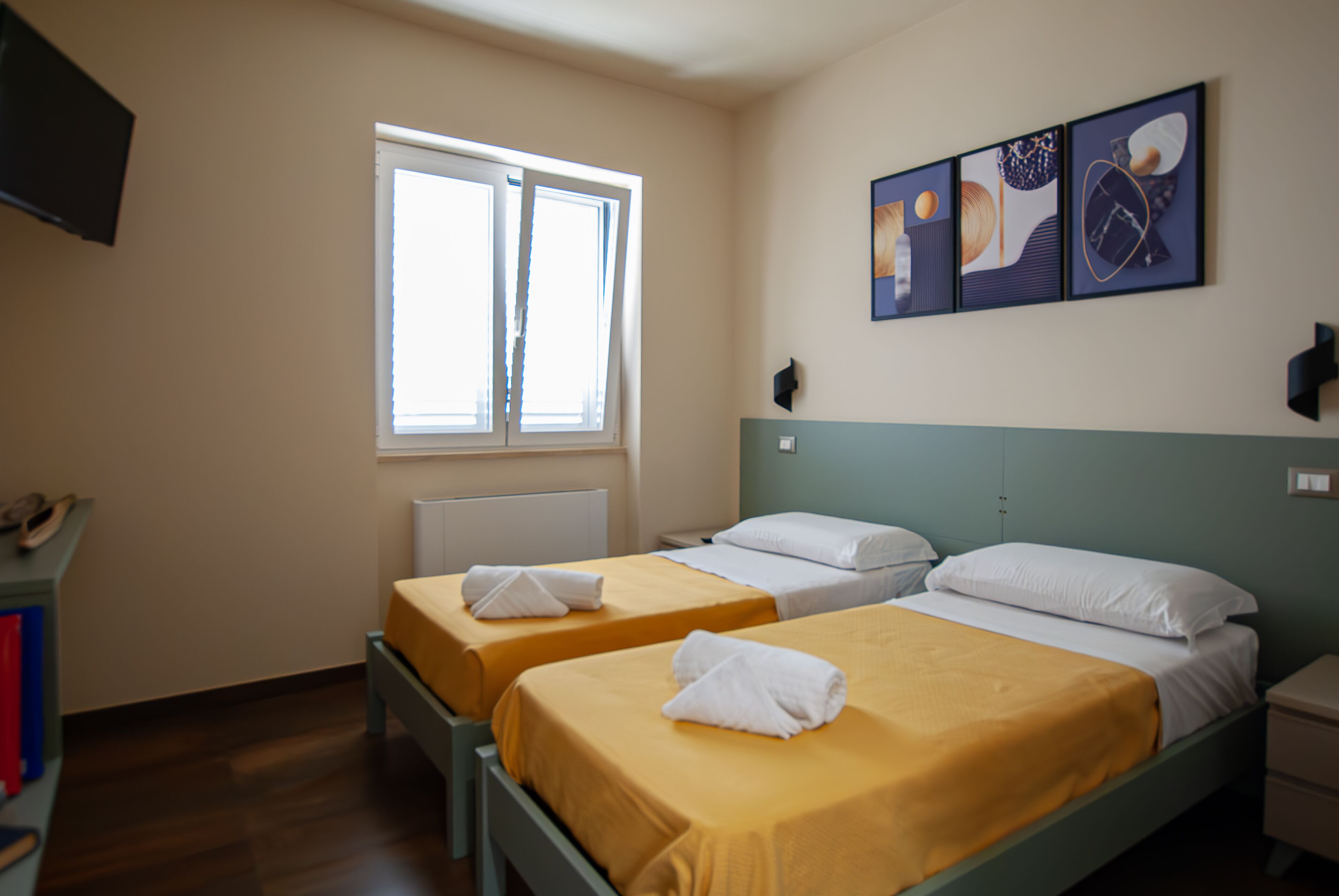 A hotel room with two single beds, a window, a radiator, a flat-screen TV, and artwork on the wall.