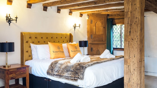 Uma cama com cabeceira estofada, roupa de cama branca e um cobertor de pele sintética. Há duas mesas de cabeceira com luminárias, uma porta de madeira e uma janela com cortinas.