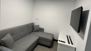 Ein moderner, minimalistischer Raum mit einem grauen Ecksofa, einem flachbildfernseher, der an der Wand befestigt ist, und einer kleinen, weißen Kommode mit einer Fernbedienung.