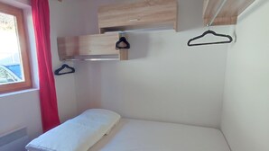 Ein kleines Zimmer mit einem Einzelbett, einem kleinen Holzregal und einem Fenster mit rotem Vorhang.