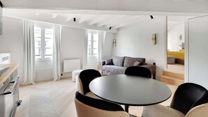 A modern living room with a sofa, a round table, and chairs. There is a kitchen area with a stove and cabinets. The room has large windows with curtains and a view of the outside. - A Refined 1-BR appartment in Saint-Germain (Paris)