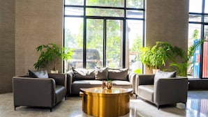A modern lobby with a sectional sofa, two armchairs, a round gold coffee table, and large windows offering a view of the street. - Sunshine Hotel (Phnom Penh)