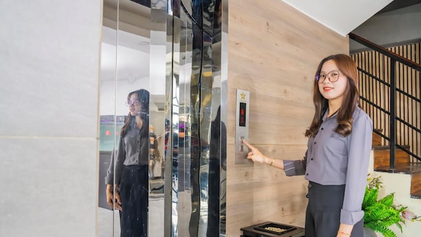 A woman standing in front of a modern elevator with a digital display, a wooden wall, and a staircase in the background. - Sunshine Hotel (Phnom Penh)