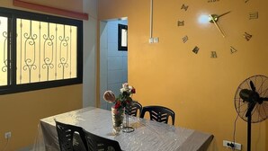 Ein Essbereich mit Tisch, der mit Folie bedeckt ist, Stühlen, einer Wanduhr, einem Ventilator und einem Fenster mit Verzierungen.