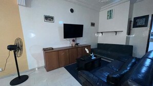 Ein Wohnzimmer mit einem schwarzen Ledersofa, einem Holzkabinett, einem an der Wand montierten Flachbildfernseher und einem Standventilator.
