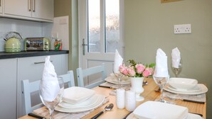 Un comedor con una mesa dispuesta para cuatro, incluyendo platos, vasos y cubiertos. Hay una jarrón con flores rosas sobre la mesa, y una zona de cocina con una tetera y un tostador visibles al fondo.