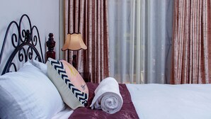 Una cama bien hecha con almohadas y una toalla enrollada, una lámpara y cortinas en una habitación de hotel.
