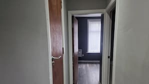 A hallway with a wooden door on the left, a white door on the right, and a window with blinds in the background. - Impeccable 3-bed Apartment in Bradford to Explore (Bradford)