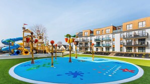 A playground with slides and climbing structures, surrounded by a pool with a blue and green design, and modern apartment buildings in the background.