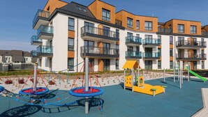 Un immeuble d’appartements moderne doté de nombreux balcons, d’une aire de jeux avec une structure d’escalade et d’une petite maison de jeu jaune.