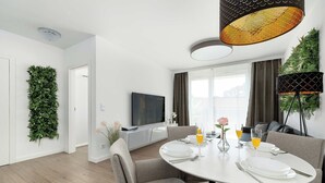 Een moderne eetruimte met een ronde tafel, gestoffeerde grijze stoelen, een flatscreen televisie en een grote plant aan de muur.