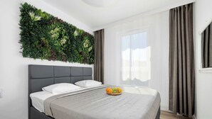 Een moderne slaapkamer met een bed, een grijs hoofdeinde, een aan de muur bevestigde plant, een raam met gordijnen, en een fruitmandje op het bed.