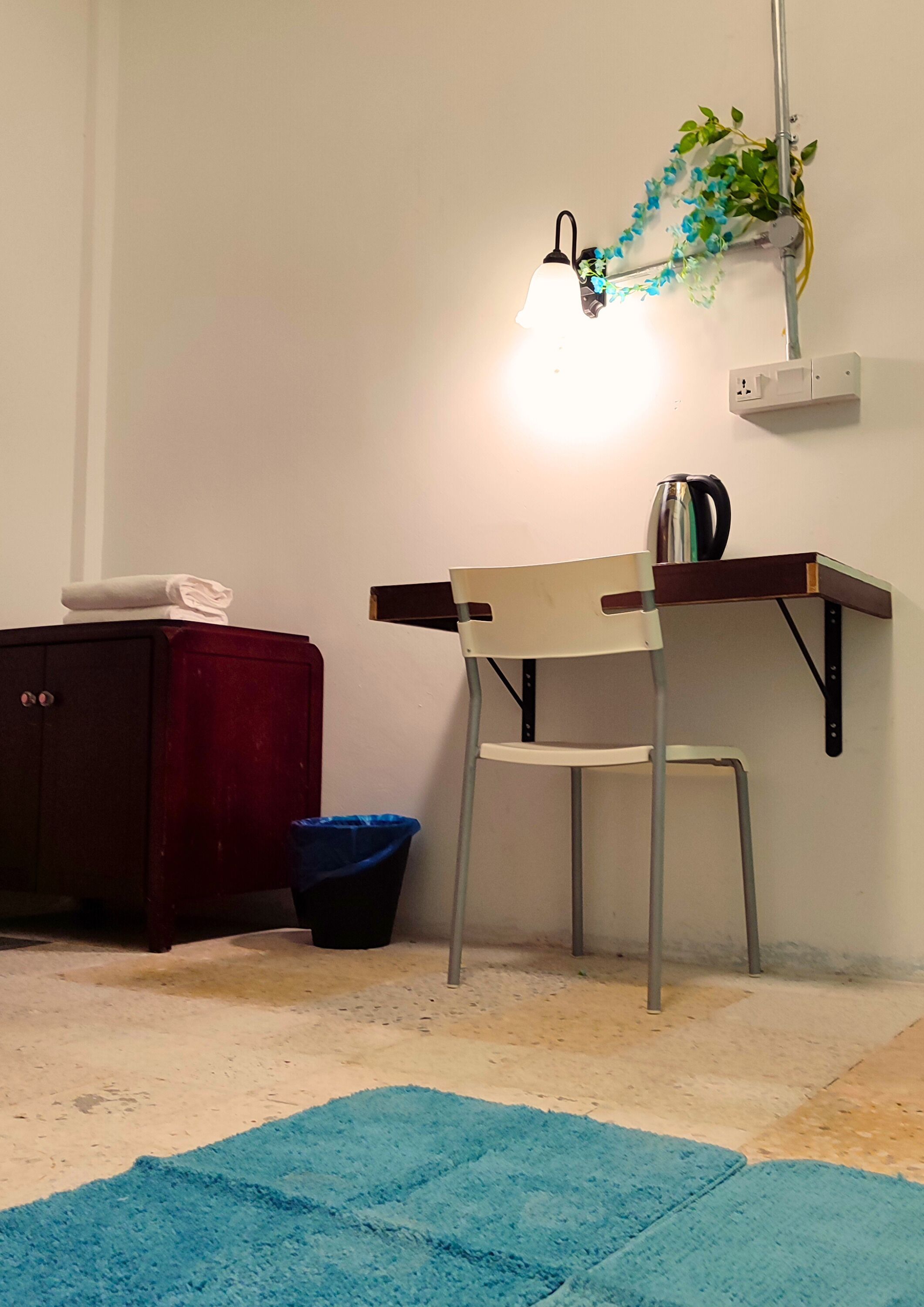 Ein kleiner Raum mit Schreibtisch, Stuhl und Lampe. Außerdem ein Papierkorb und eine Heizung an der Wand.