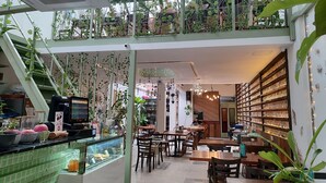 Ein Café mit einer grün gefliesten Theke, Holztischen und -stühlen sowie einem Balkon im zweiten Stock mit Pflanzen und Lichterketten.