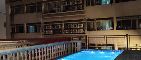A swimming pool with a tiled edge, surrounded by a railing, and a building with multiple floors and balconies in the background.
