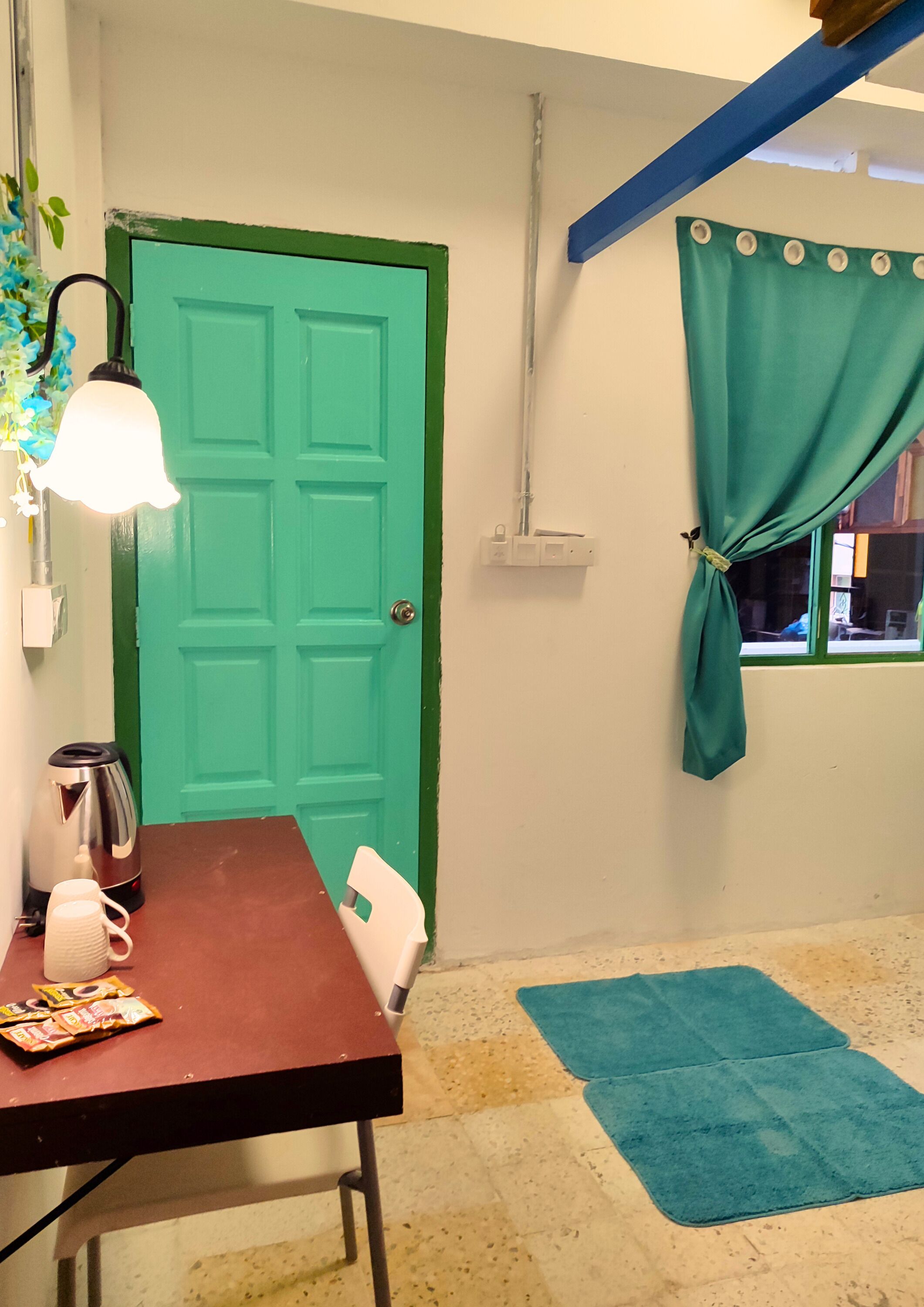 Ein Zimmer mit einer türkisfarbenen Tür, einem Tisch mit Wasserkocher und Tassen, einem Stuhl, einem blauen Teppich und einem Fenster mit zurückgebundenen türkis-farbenen Vorhängen.