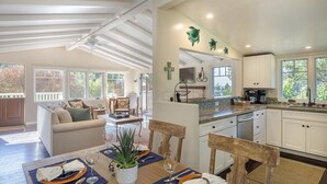 A spacious kitchen with white cabinets, a dining area with a wooden table and chairs, and a cozy living room with a sofa and a large window.