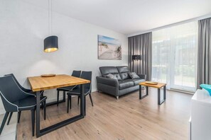 Uma sala de estar moderna com uma mesa de jantar de madeira, cadeiras pretas, um sofá cinzento e uma janela grande com cortinas.