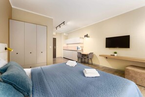 A hotel room with a bed, a desk, a chair, a TV, and a kitchenette.