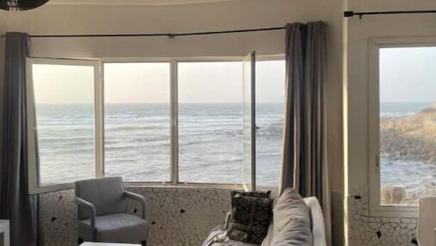 Ein modernes Wohnzimmer mit Sofa, Sessel und Couchtisch. Große Fenster bieten Blick auf das Meer.