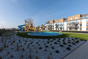 Un parque infantil con toboganes y estructuras para escalar, rodeado de un área ajardinada con grava y césped.