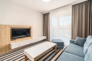 A modern living room with a flat-screen TV, a white coffee table, a blue sofa, and a large window with curtains.