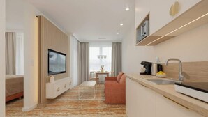 A modern hotel room with a TV, kitchenette, and a seating area with a sofa and table.