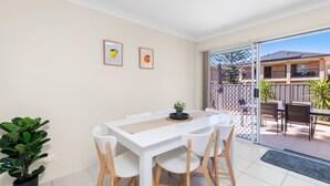 Un comedor con una mesa y sillas blancas, dos cuadros en la pared y un ventanal corredizo que da a un patio exterior con cerramiento y plantas.