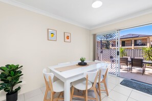 Ein Essbereich mit einem weißen Tisch und Stühlen, zwei gerahmten Bildern an der Wand und einer Glasschiebetür, die auf eine Außenpatio mit Zaun und Pflanzen führt.