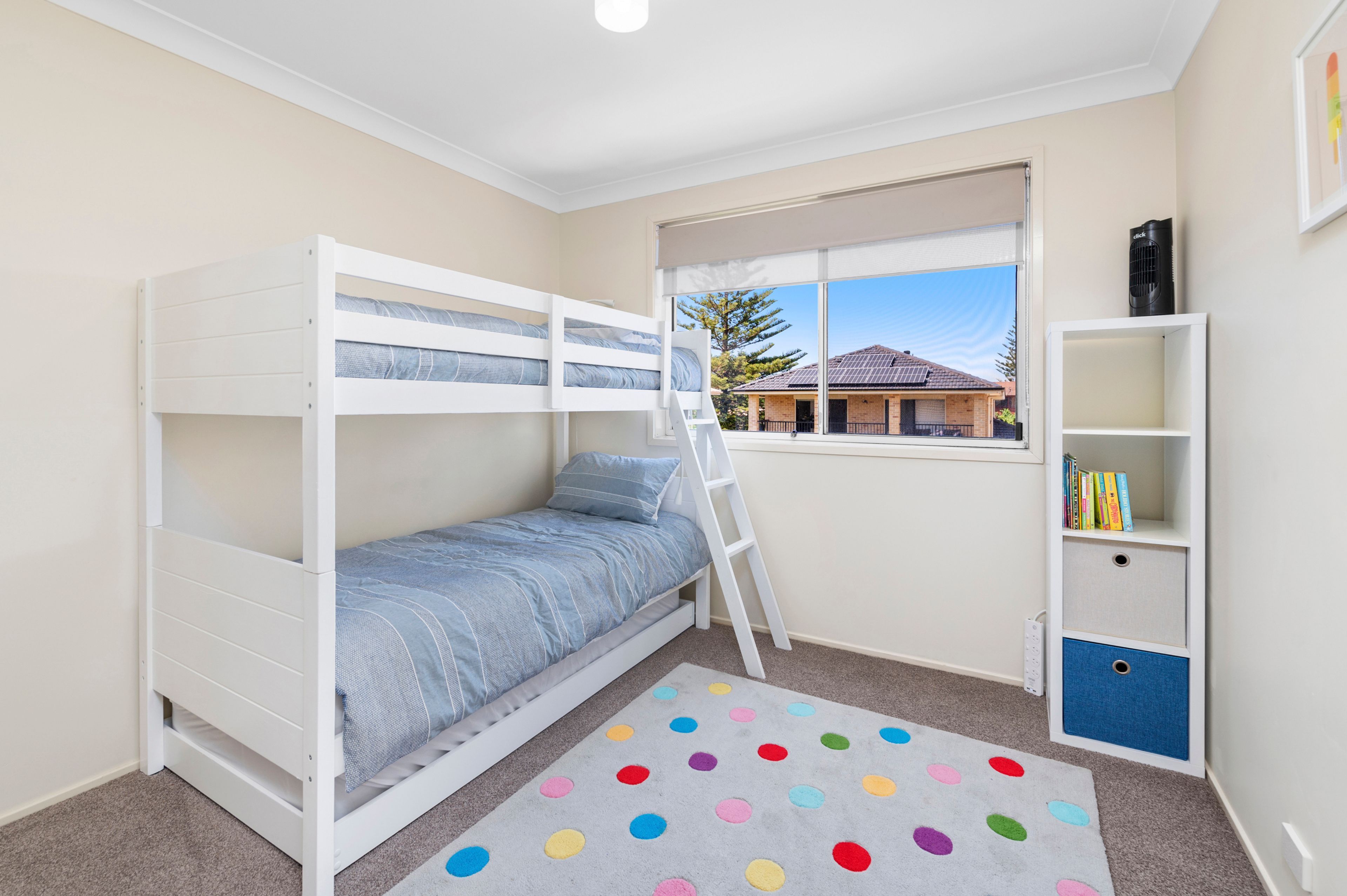 Un dormitorio infantil con literas, una ventana con vistas, una estantería y una alfombra de lunares.