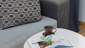 A small round table with a magazine and a cup, placed on a patterned rug in a living room with a grey sofa and patterned pillows.