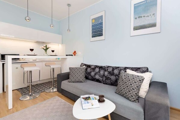 A modern living space with a kitchen, a grey sofa with patterned cushions, a small white table, and pendant lighting.