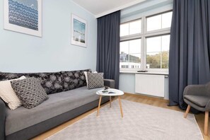 A modern living room with a grey sofa, patterned cushions, a small white table, and a view through large windows with dark curtains.