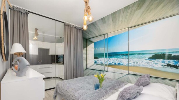 A modern bedroom with a large bed, a bedside table, a mirror, and a view of the ocean through a large window. - Stylish Apartment with Balcony | Parking | Close to the Beach (Kolobrzeg)