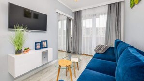 A modern living room with a blue sofa, a glass coffee table, a white console table with plants, and a flat-screen TV mounted on the wall. - Stylish Apartment with Balcony | Parking | Close to the Beach (Kolobrzeg)
