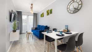 A modern living room with a blue sofa, a dining table with chairs, a wall-mounted clock, and a flat-screen TV. - Stylish Apartment with Balcony | Parking | Close to the Beach (Kolobrzeg)