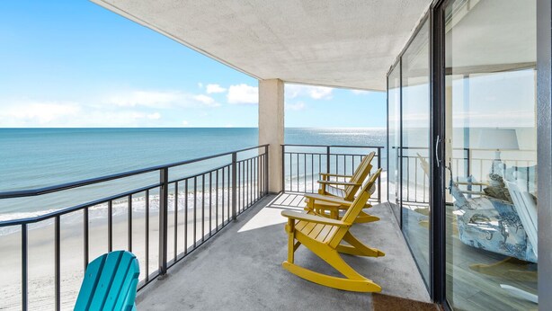A balcony with a view of the ocean, two rocking chairs, and a glass door leading to a room with a lamp and decorative items. - Condo with Private Balcony & Beach Views Near Pier (Garden City Beach)