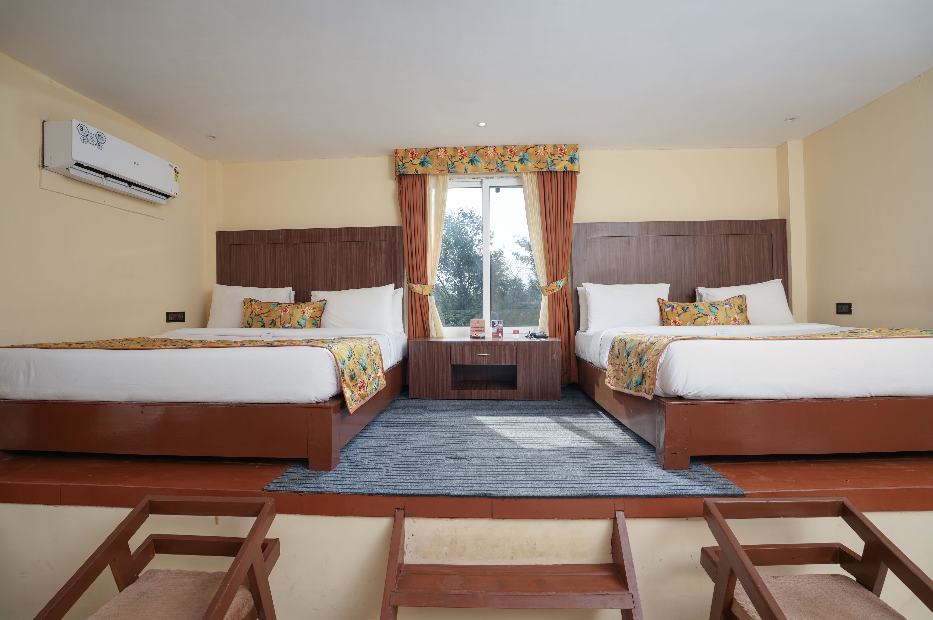 A hotel room with two beds, a window with curtains, an air conditioning unit, and a small table between the beds.