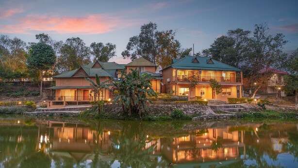 Un'ampia casa a più piani con uno stagno riflettente di fronte, circondata da alberi e un cielo al tramonto.