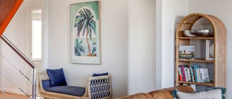 En stue med en fletstol, en lædersofa, en reol og et billede af palmer.