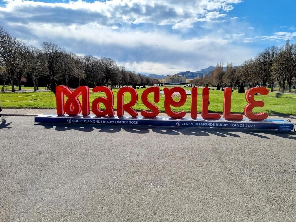 Un grand panneau rouge sur lequel on peut lire le mot « MARSEILLE », avec un livre intitulé « COUPE DU MONDE DE RUGBY, FRANCE 2023 » placé en dessous.