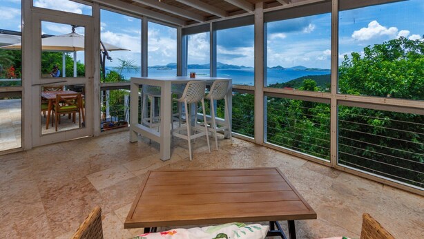 Een overdekt terras met een houten tafel en rieten stoelen, met uitzicht op zee en groen.